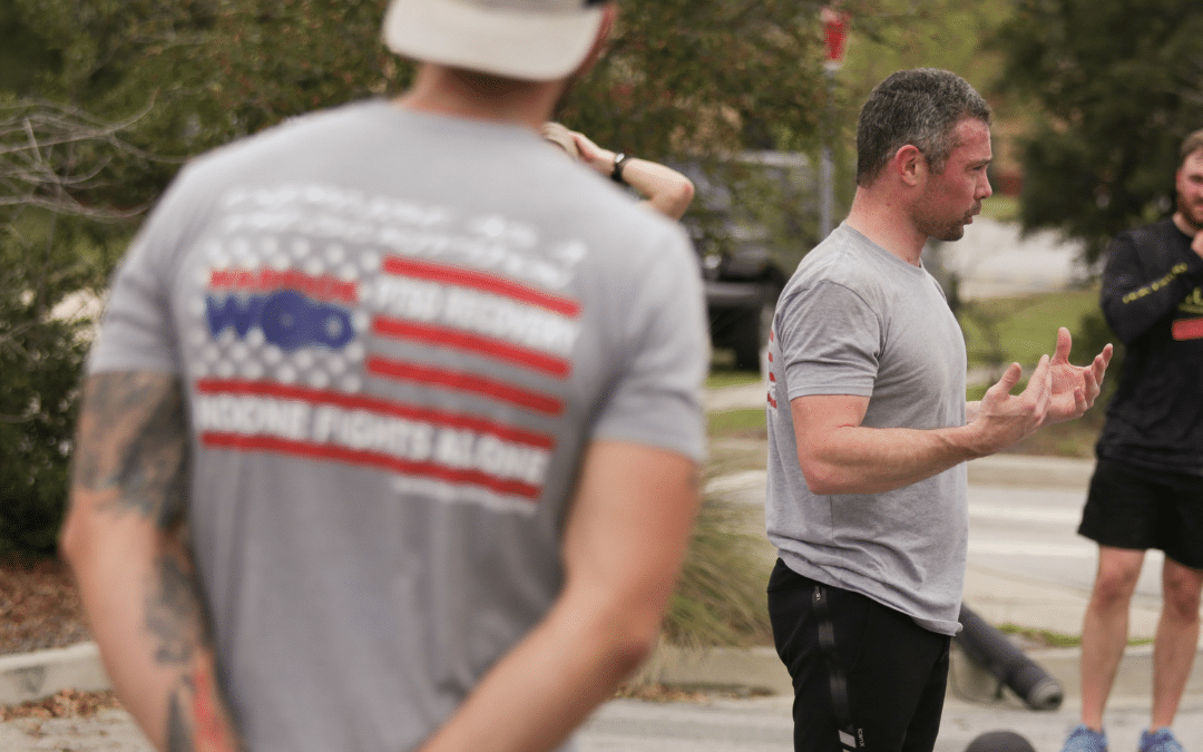 Group of veterans participating in an outdoor WarriorWOD exercise session, with one person addressing the group and another observing with arms crossed, wearing WarriorWOD shirts.