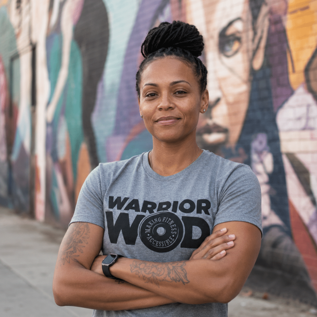Woman Army veteran standing with arms crossed in front of a mural, wearing a grey WarriorWOD t-shirt.