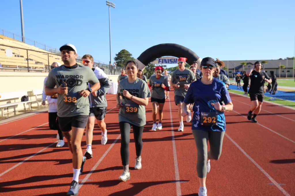 A group of Hustle for Heroes 5K participants in WarriorWOD shirts running together on the track past the finish arch