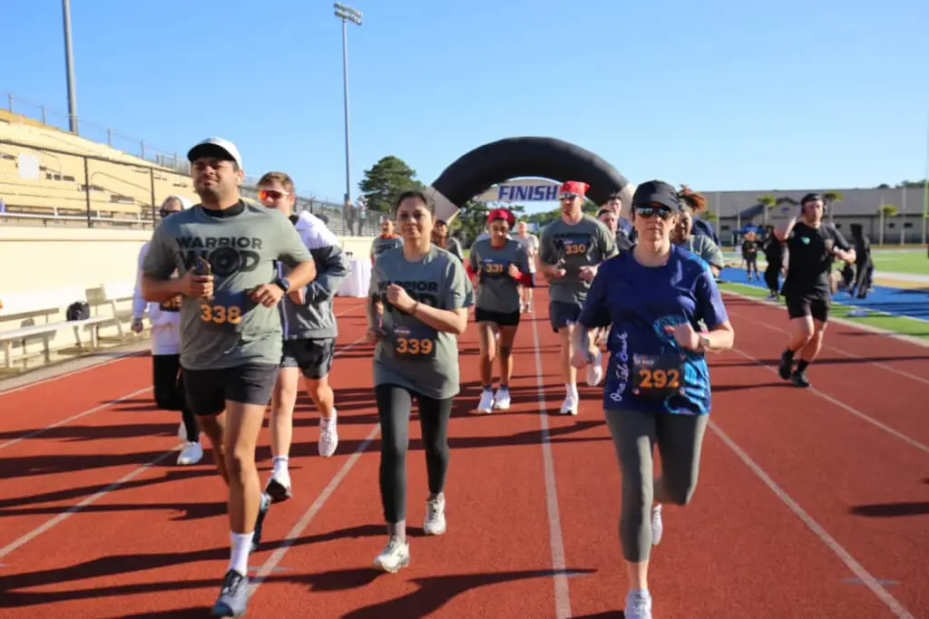 A group of Hustle for Heroes 5K participants in WarriorWOD shirts running together on the track past the finish arch