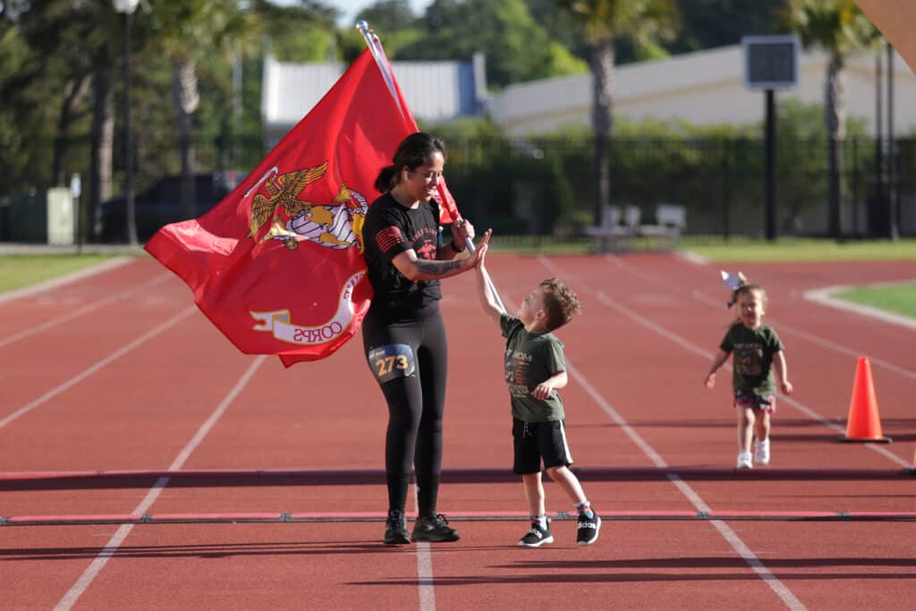 A woman carrying the Marine Corps flag high-fives her young son on the track at the Hustle for Heroes 5K while a toddler runs behind them