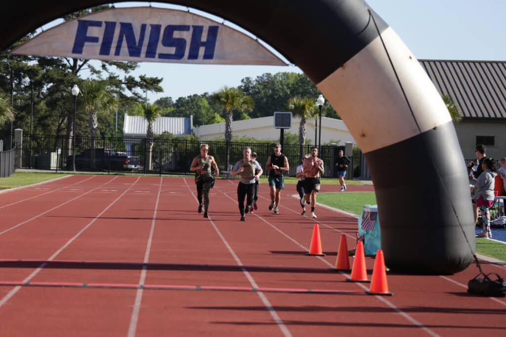 Runners and a rucker approach the finish arch at the Hustle for Heroes 5K on the track at Buccaneer Field