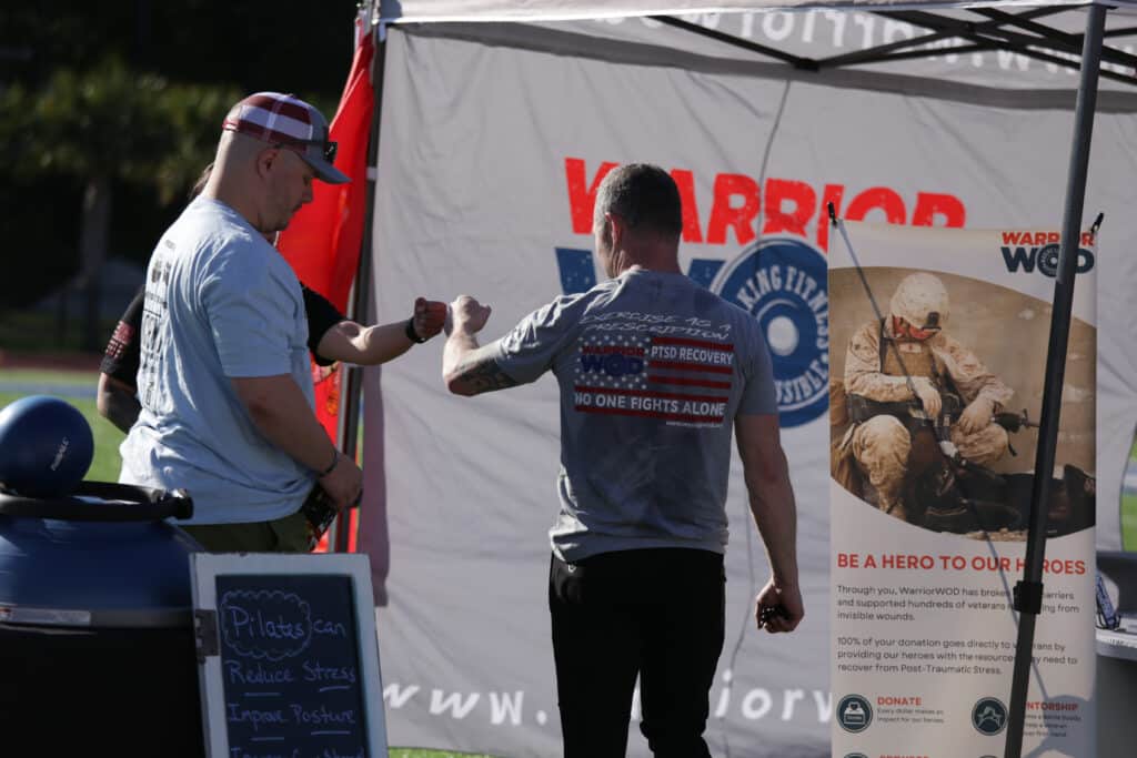 Two men fist bump at the WarriorWOD tent at the Hustle for Heroes 5K, in front of a banner reading No One Fights Alone