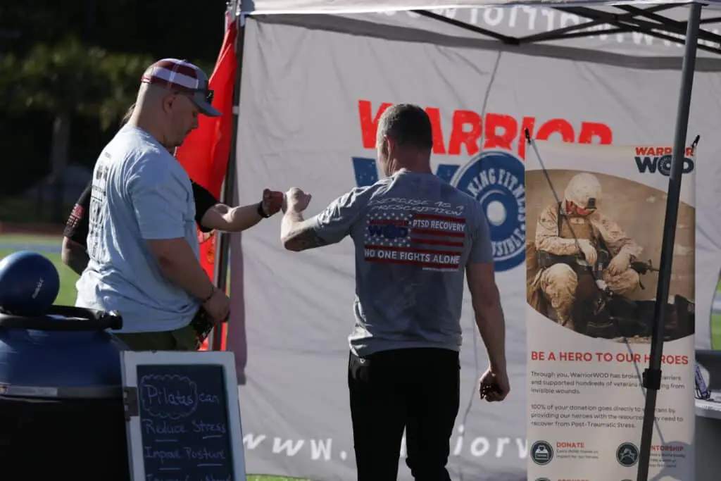 Two men fist bump at the WarriorWOD tent at the Hustle for Heroes 5K, in front of a banner reading No One Fights Alone