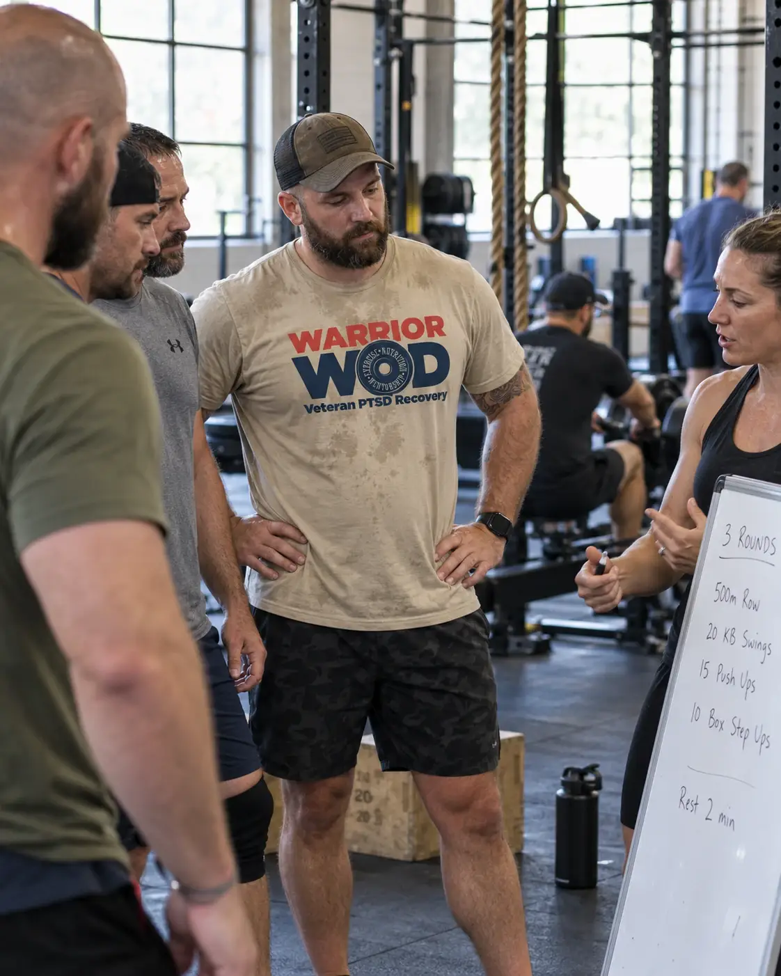 Veterans in WarriorWOD shirts listen as a coach briefs a 3-round workout on a whiteboard at a Preferred Partner Gym