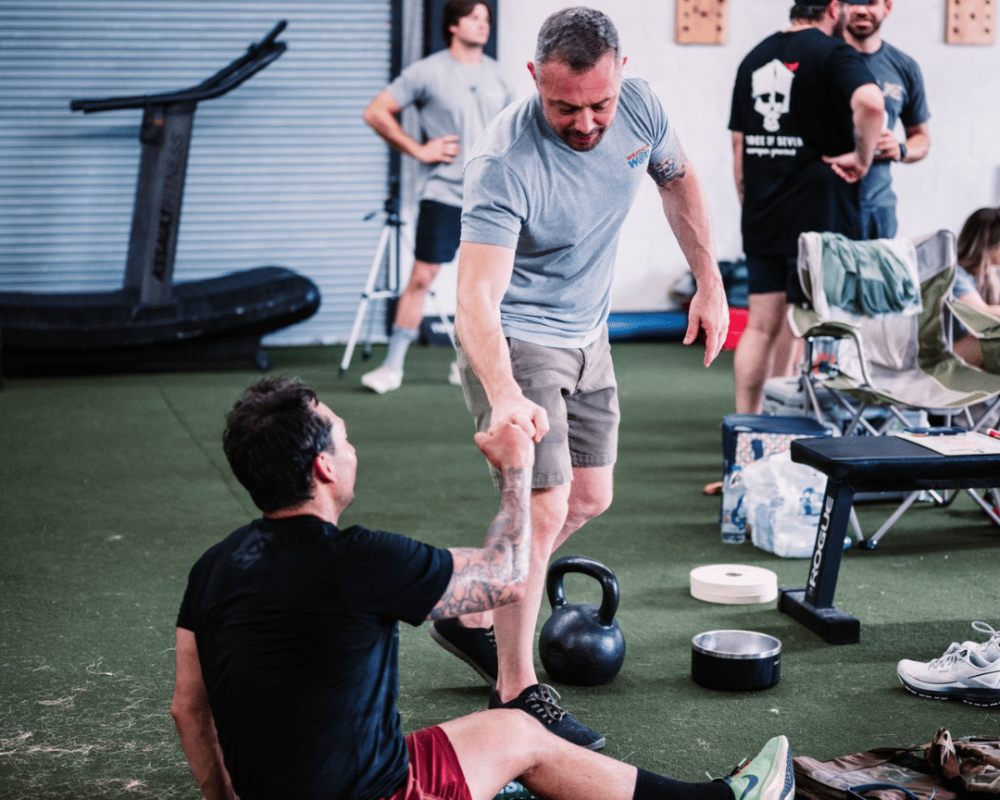 A WarriorWOD veteran reaches down to help a fellow Warrior up off the gym floor after a workout, kettlebell and weights nearby.