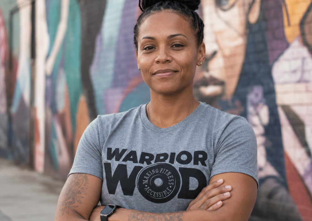 Woman Army veteran standing with arms crossed in front of a mural, wearing a grey WarriorWOD t-shirt.